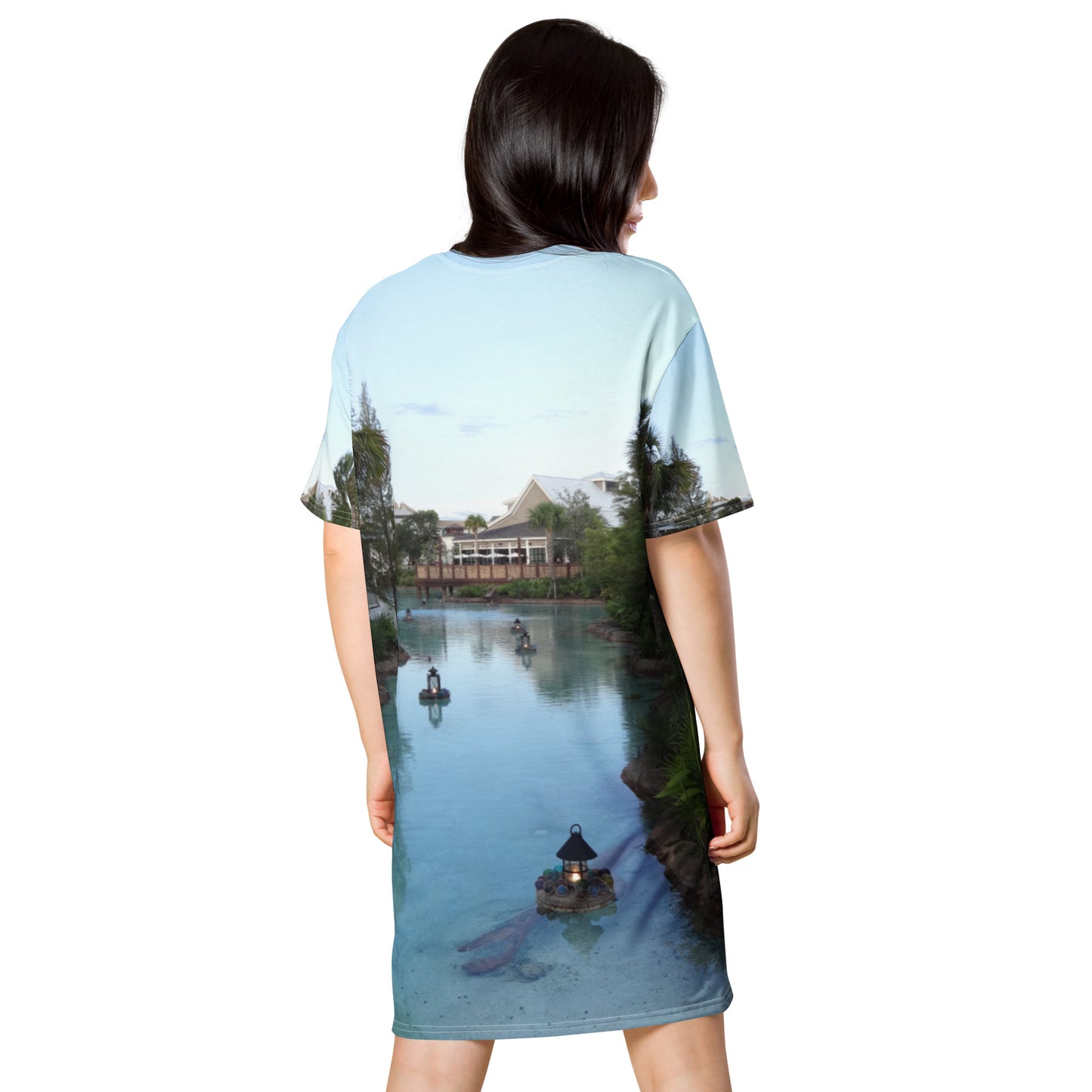 Pictured is a women wearing an all over print tshirt dress with a photograph of a little river flowing through a village - dress back