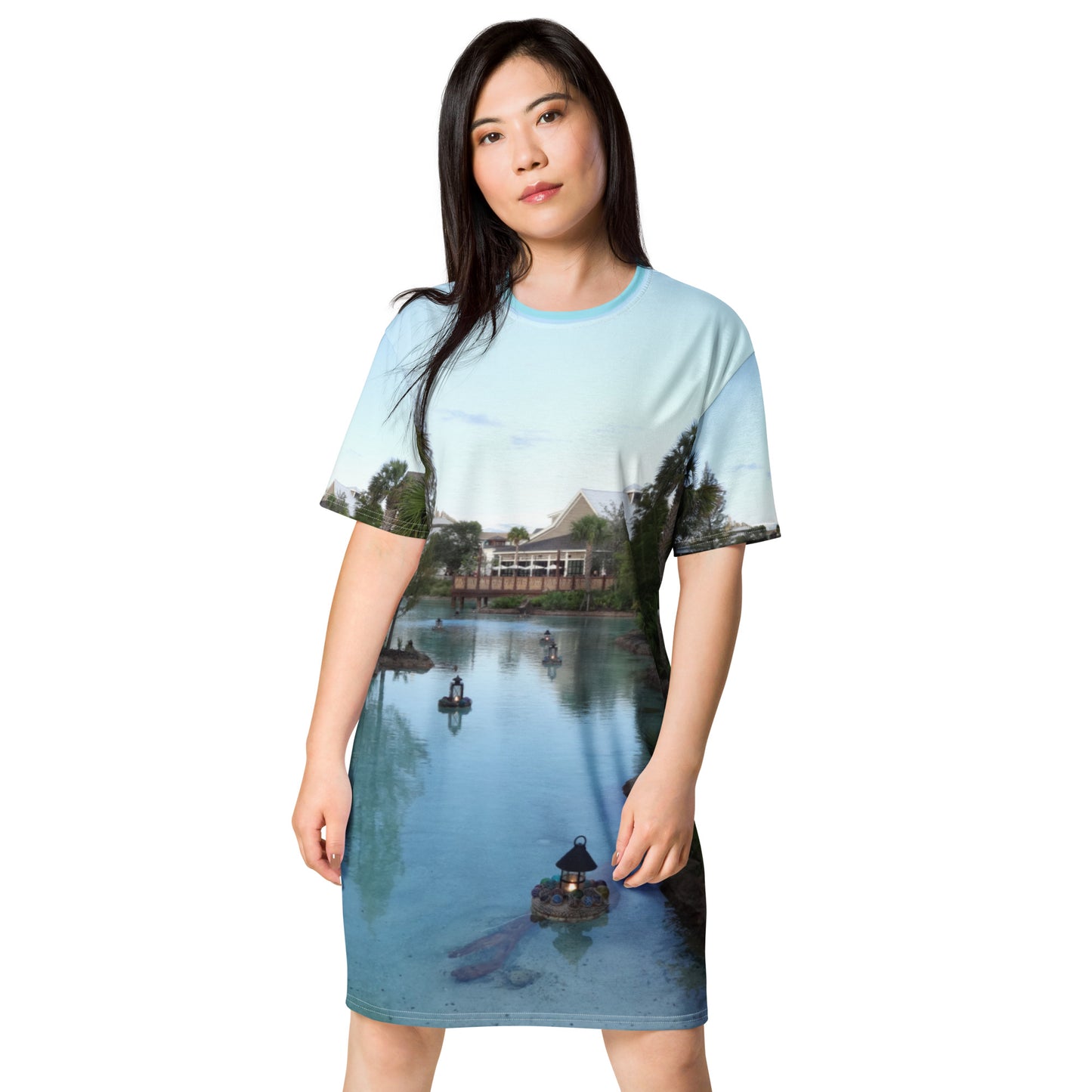 Pictured is a women wearing an all over print tshirt dress with a photograph of a little river flowing through a village - dress front
