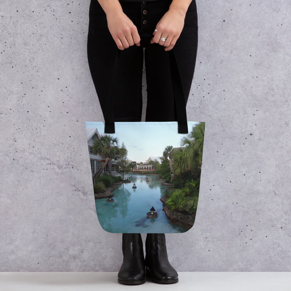 Pictured is a person holding a all over print tote bag with a photograph of a little river flowing through a village - black handles