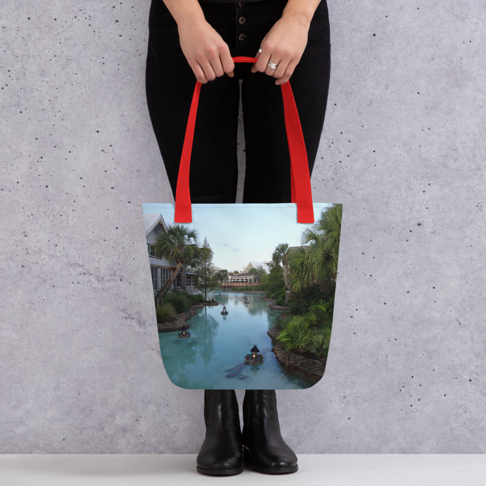 Pictured is a person holding a all over print tote bag with a photograph of a little river flowing through a village - red handles