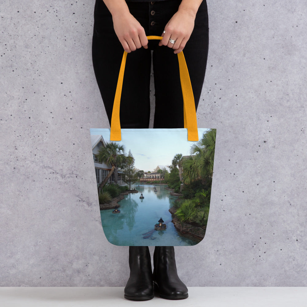 Pictured is a person holding a all over print tote bag with a photograph of a little river flowing through a village - yellow handles