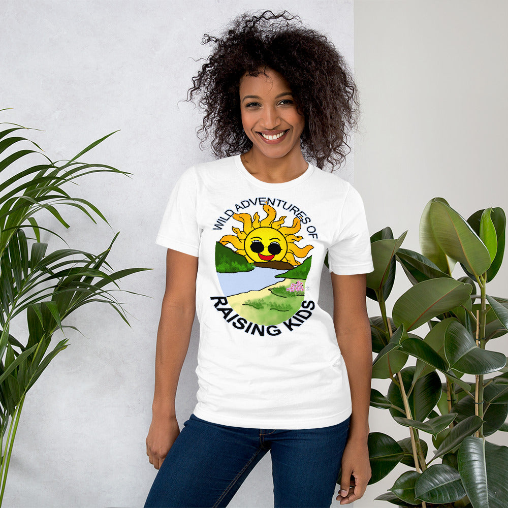 A woman wearing a tshirts with a picture of the sun coming up over the mountains with sunglasses on and a little stream gently flowing trees and bushes can be seen on the closer mountains and a small beach with some flowers are on the closes bank around the outside of the picture is written "Wild Adventures of Raising Kids" unisex tshirt - white front